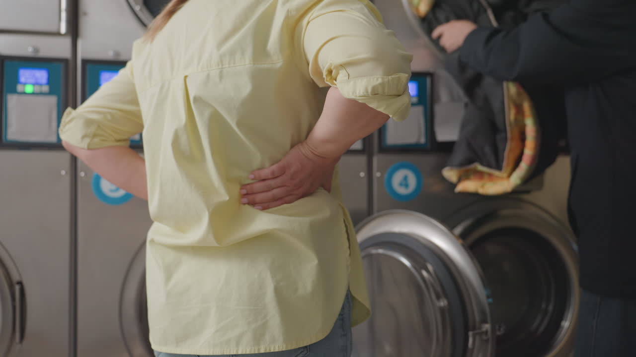 Back view business woman with hands on waist watching man unload clothes from washer inside laundromat, stainless machines in row, professional service workflow, indoor hygiene operation