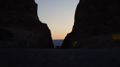 Coche-Conduciendo-Por-Una-Carretera-Desértica-A-Través-De-Dos-Grandes-Laderas-De-Montañas-Al-Atardecer