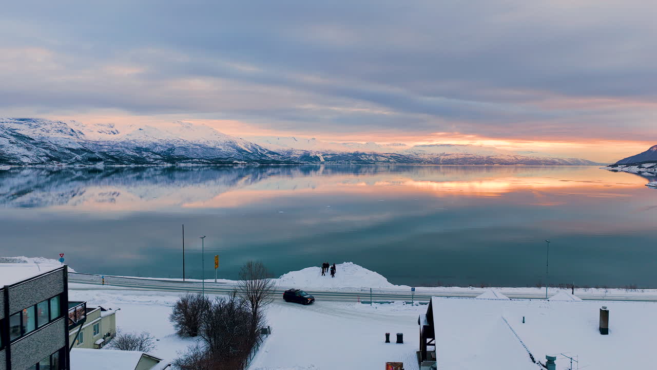 ノルウェーのロフォーテン諸島にある穏やかなフィヨルドの息をのむような空撮。冬の夕焼けに染まり、雪に覆われた山々と村の家々が広がる