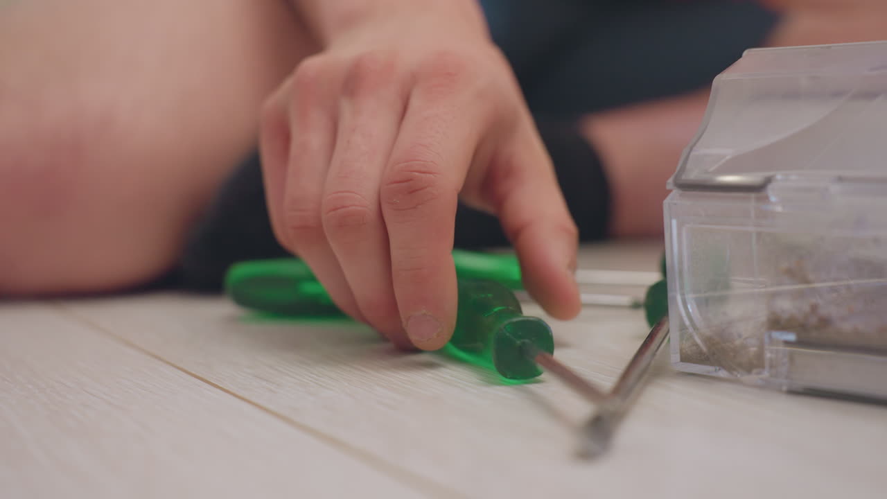 Close up of engineer hand picking up screw beside green tools and dusty robot vacuum container while seated on floor during repair process with focus on maintenance