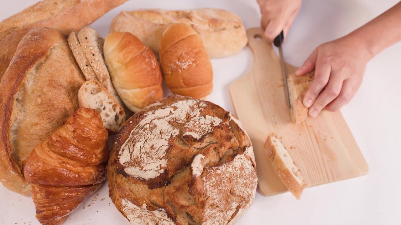 Close-up overhead shot of hands slicing bread beside sourdough, croissants, and various rustic loaves.