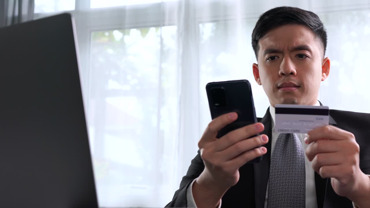Pengusaha muda Asia yang cerdas duduk di kantor rumah dengan laptop, tersenyum dan bahagia saat menggunakan kartu kredit dengan aplikasi belanja online di ponsel pintar