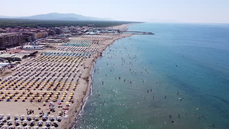 Wunderschöne-Luftaufnahme-Eines-Sandstrandes-Voller-Sonnenschirme-Mit-Menschen,-Die-Am-Meer-Schwimmen,-Und-Einem-Fernen-Berg-Im-Hintergrund