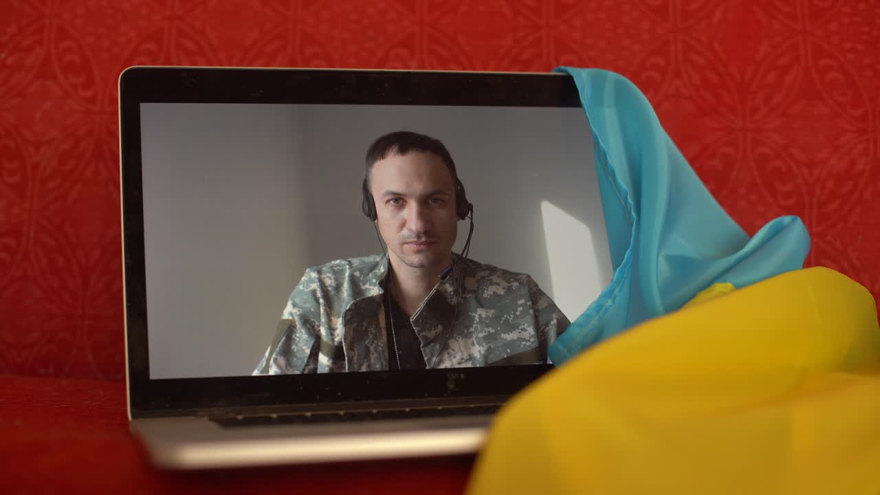 Soldato in uniforme militare siede sul divano di casa usando un laptop con webcam per comunicare con la famiglia. Bandiera ucraina sullo sfondo. Azioni militari in Ucraina. Popolo libero e democratico nello stato