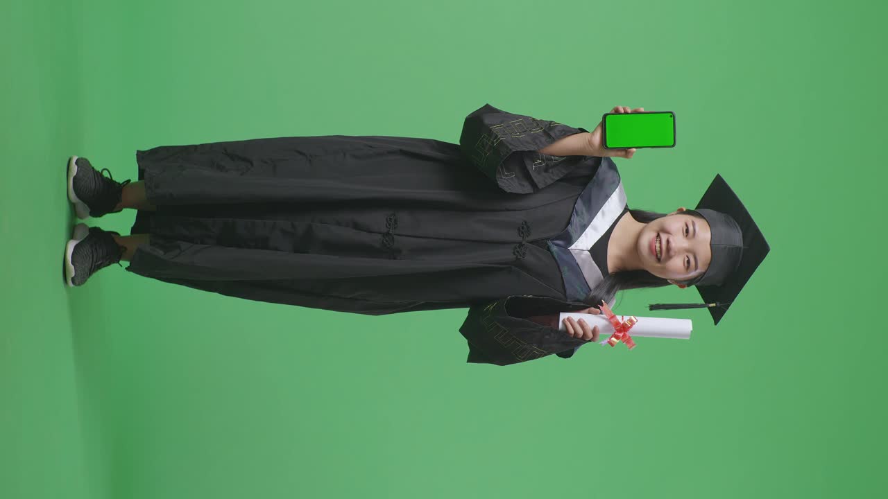 Studentessa asiatica laureata a figura intera, con tocco e toga, che sorride e mostra il diploma e uno smartphone con schermo verde alla telecamera, su sfondo a schermo verde in studio