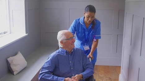 senior man at home being pushed in wheelchair by female care worker in uniform