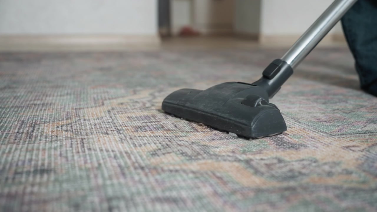 Hlavice vysavače na koberce klouzající po vzorovaném koberci, asijská uklízečka se soustředí na vlákna a sání, detail trysky, texturovaný povrch podlahy, přesný pohyb a profesionální atmosféra úklidu domácnosti.