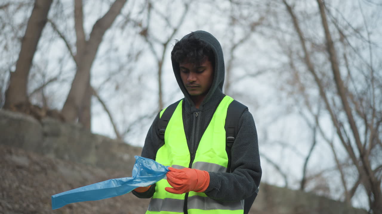 Young man in reflective safety vest and orange gloves preparing blue trash bag during outdoor park cleanup in cold weather, focused expression while volunteering for environmental conservation activity