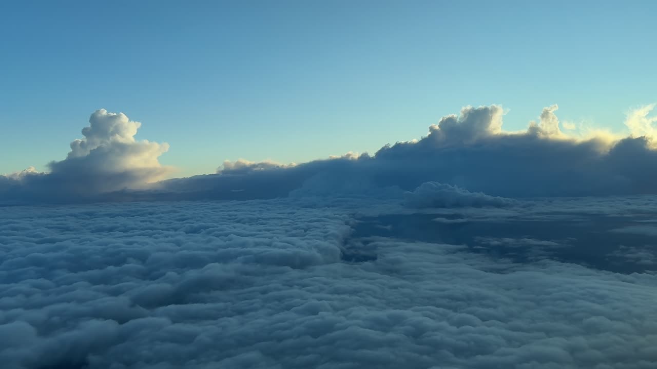 A pilot perspective POV from a jet airplane cockpit while flying at sunset over an endless sea of clouds and with threatening storm clouds ahead.