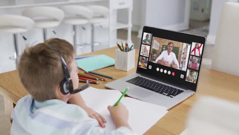 Schoolboy-using-laptop-for-online-lesson-at-home,-with-diverse-teacher-and-class-on-screen