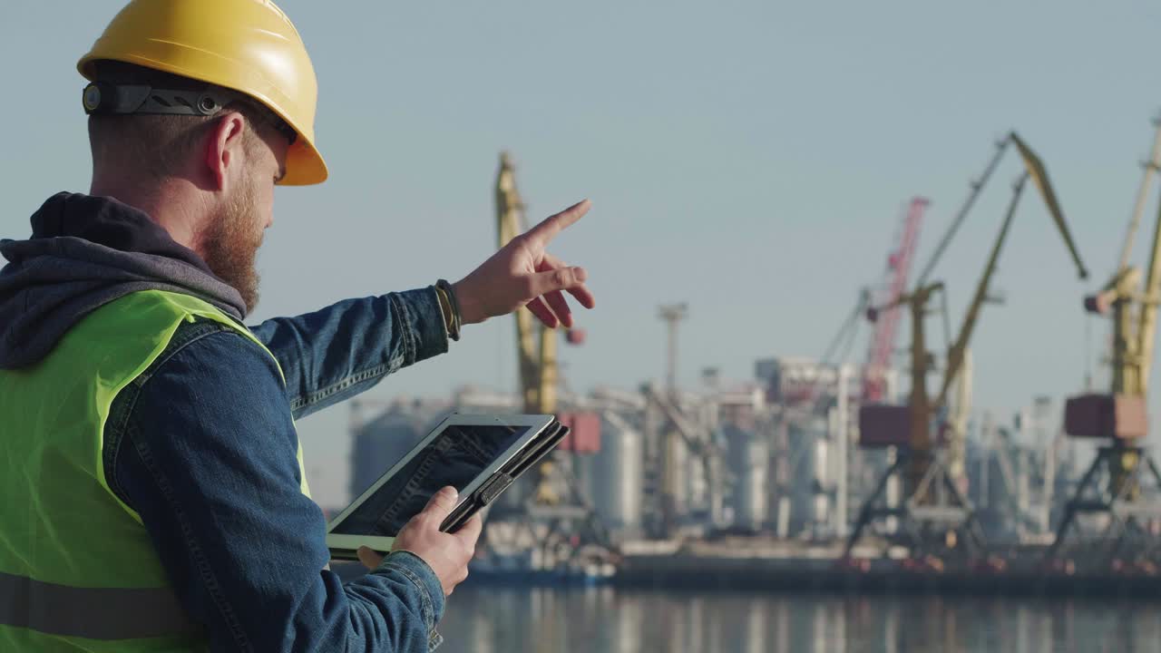 un trabajador portuario con barba en un casco amarillo, trabaja con una tableta pc en el puerto contra el fondo de grúas. el capataz inspecciona el puerto industrial. cine 4k video