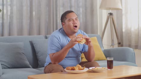 a fat asian man smelling to hamburger in his hands then smiling while eating fast food on a sofa in the living room at home