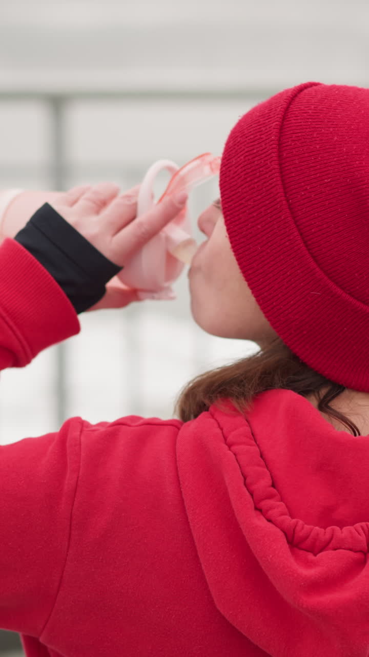 lady in red beanie and hoodie sipping water from pink bottle outdoors drops bottle, blurred background of iron rail in serene winter setting with light snow and calm atmosphere