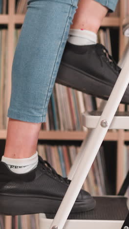 woman goes up step ladder near bookcase closeup. librarian uses foldable stairs to take books from upper shelf in library. student works in bookstore