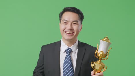 close up of asian business man in a suit and tie with a gold medal and trophy showing peace gesture and smiling to camera as the first winner on green screen background in the studio