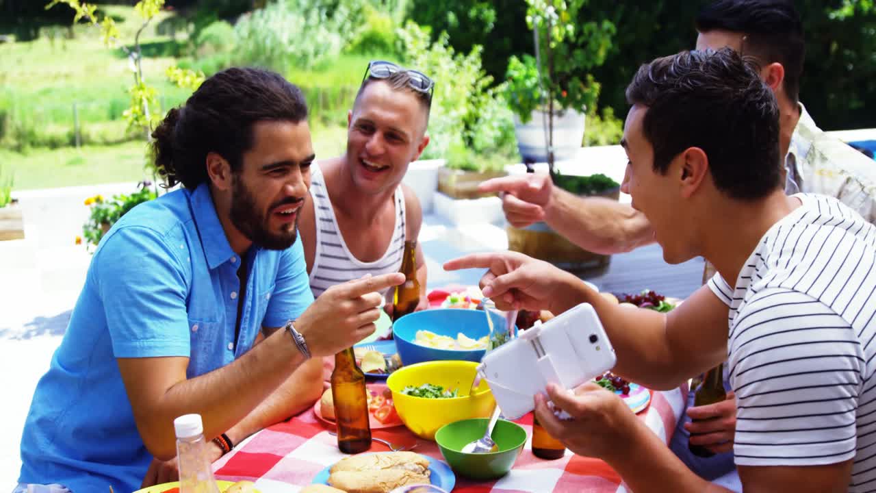 hombre sonriente mostrando su teléfono móvil a sus amigos mientras come al aire libre