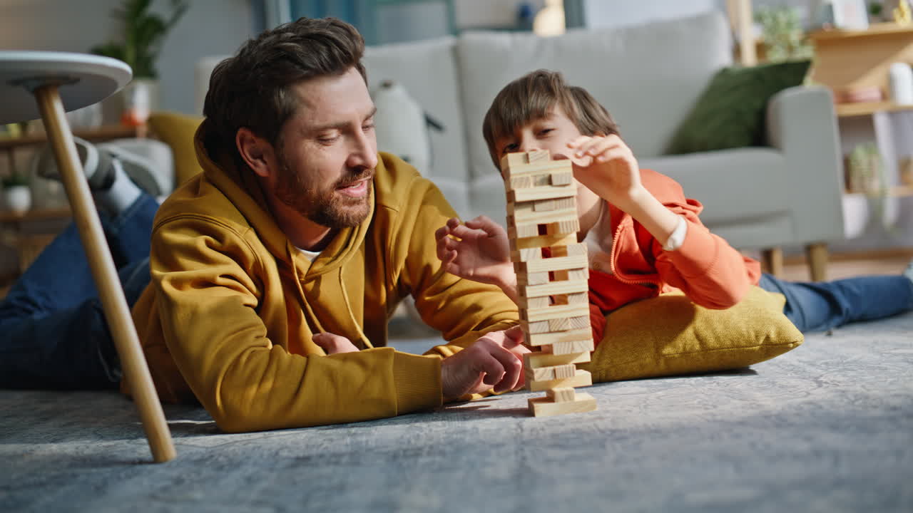 Papà e bambino si divertono con il Jenga sdraiati sul pavimento, in primo piano. Un figlio giocoso costruisce una torre di giocattoli.