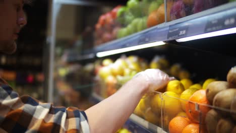 Close-up-of-shop-assistants-in-transparent-gloves-filling-up-lemons-storage-stands-with-assorted-organic-groceries-in-supermarket.-Supermarket-clerks-placing-fruits-on-display-store-shelves