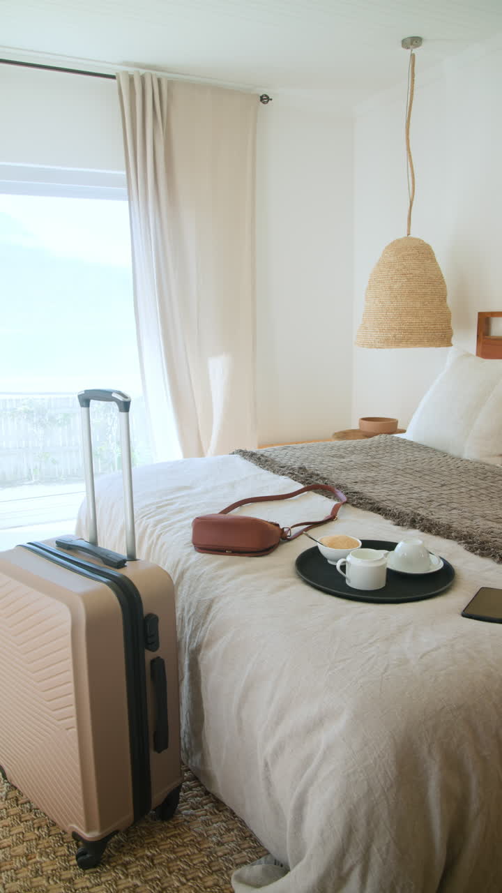 Video vertical: Mujer viajera relajándose en una habitación de hotel con maleta, café y smartphone.