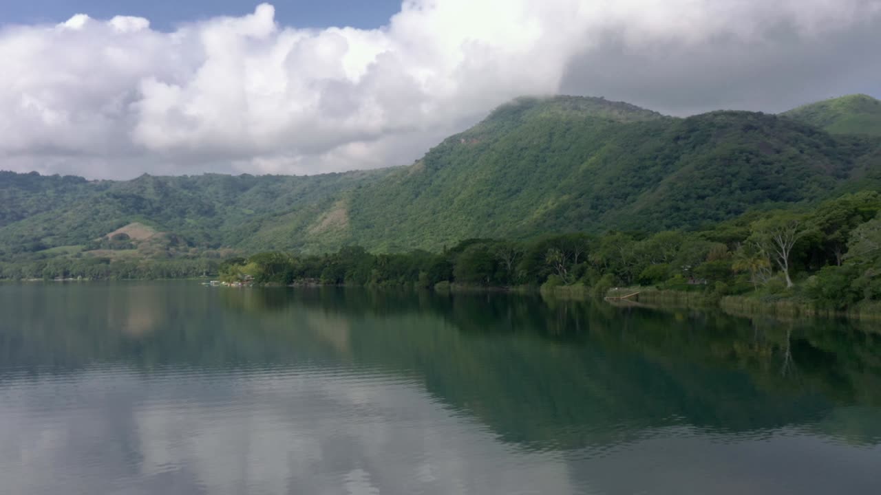 Serene static shot of a mountain lake reflecting clouds in its calm water, flanked by lush forested hills under soft, diffused lighting. No people present for tranquil mood