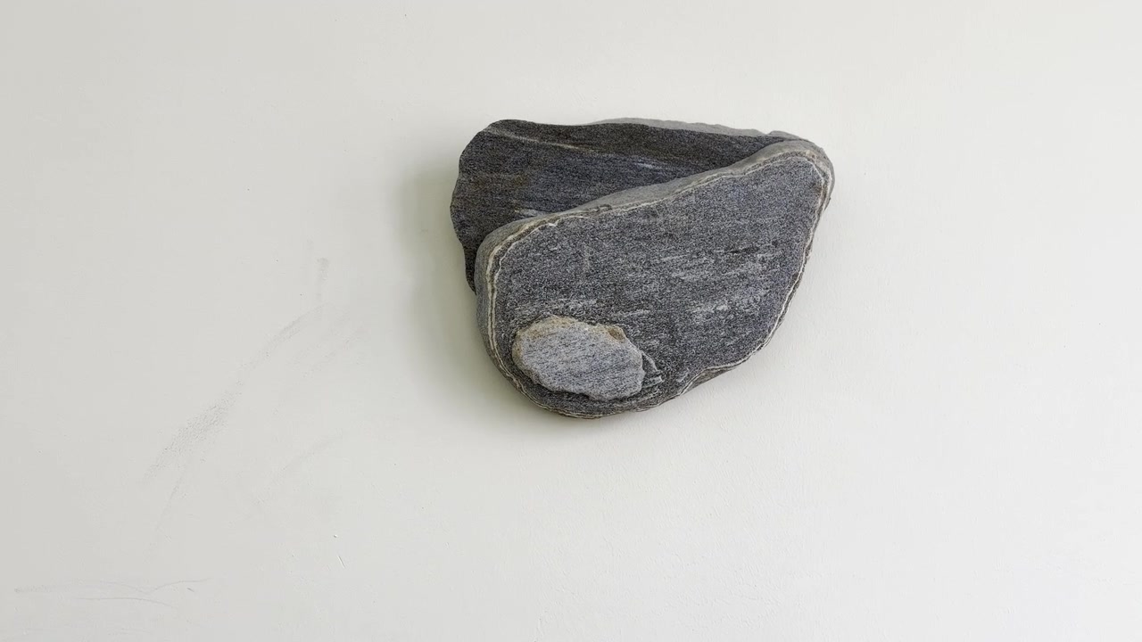Ein dunkelgrauer Stein hängt als Dekoration an einer hellen Wand; er hat einen hellen Fleck darauf. Nahaufnahme.