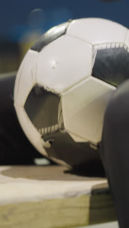 primer plano de un hombre sentado en un banco con una pelota de fútbol sostenida entre sus piernas, está tocando su pie en el suelo repetidamente con un fondo borroso