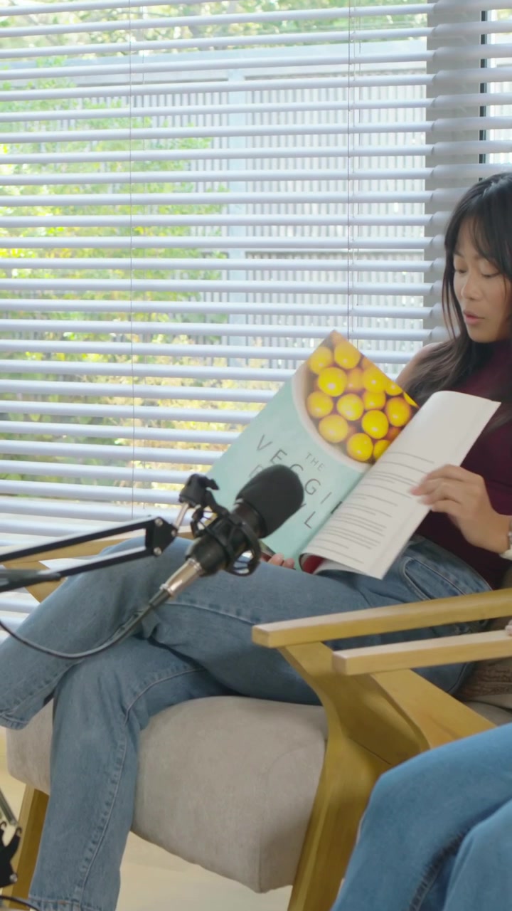 Video verticale: Donna con top bordeaux che sfoglia una rivista dopo che il co-conduttore le fa un cenno per il podcast.