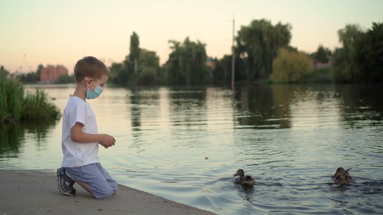 의료 마스크를 입은 소년이 호수 01에서 오리에게 먹이를 주고 있다.