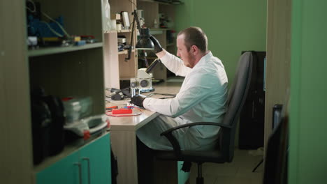 una vista de cerca de un técnico con una bata de laboratorio blanca y guantes de mano negros, ajustando una lámpara de escritorio, el técnico luego continúa trabajando bajo el microscopio, con varias herramientas alrededor