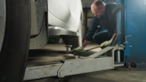 focused view on a car jack being positioned under a vehicle in a workshop, with a mechanic's presence suggested by visible gloved hands and a blue lift pole