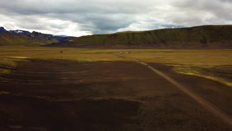 Vista-Aérea-Del-Paisaje-Sobre-Unos-Pocos-Vehículos-Que-Circulan-A-Toda-Velocidad-Por-Un-Camino-De-Tierra-A-Través-De-Las-Tierras-Altas-Islandesas
