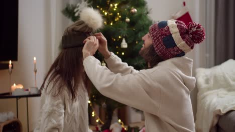 A-happy-guy-in-a-New-Year's-warm-hat-puts-a-green-brown-hat-on-the-head-of-his-brunette-girlfriend-in-a-White-sweater.-They-look-at-each-other-and-smile-near-the-New-Year-tree-in-the-atmosphere-of-Christmas-and-New-Year-at-home-in-the-evening