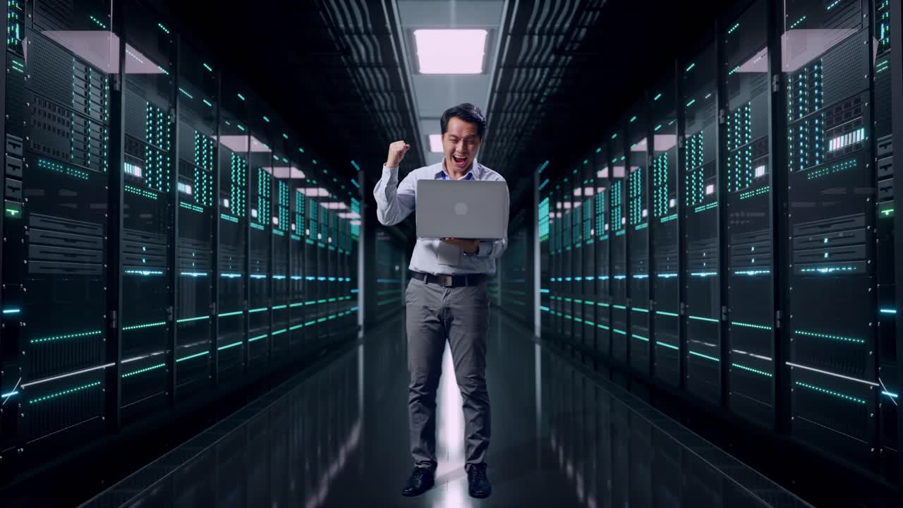 Helkropsbillede af en asiatisk professionel mandlig medarbejder, der står med sin bærbare computer i et serverrum i et datacenter, og hæver knyttet næve i jubel.