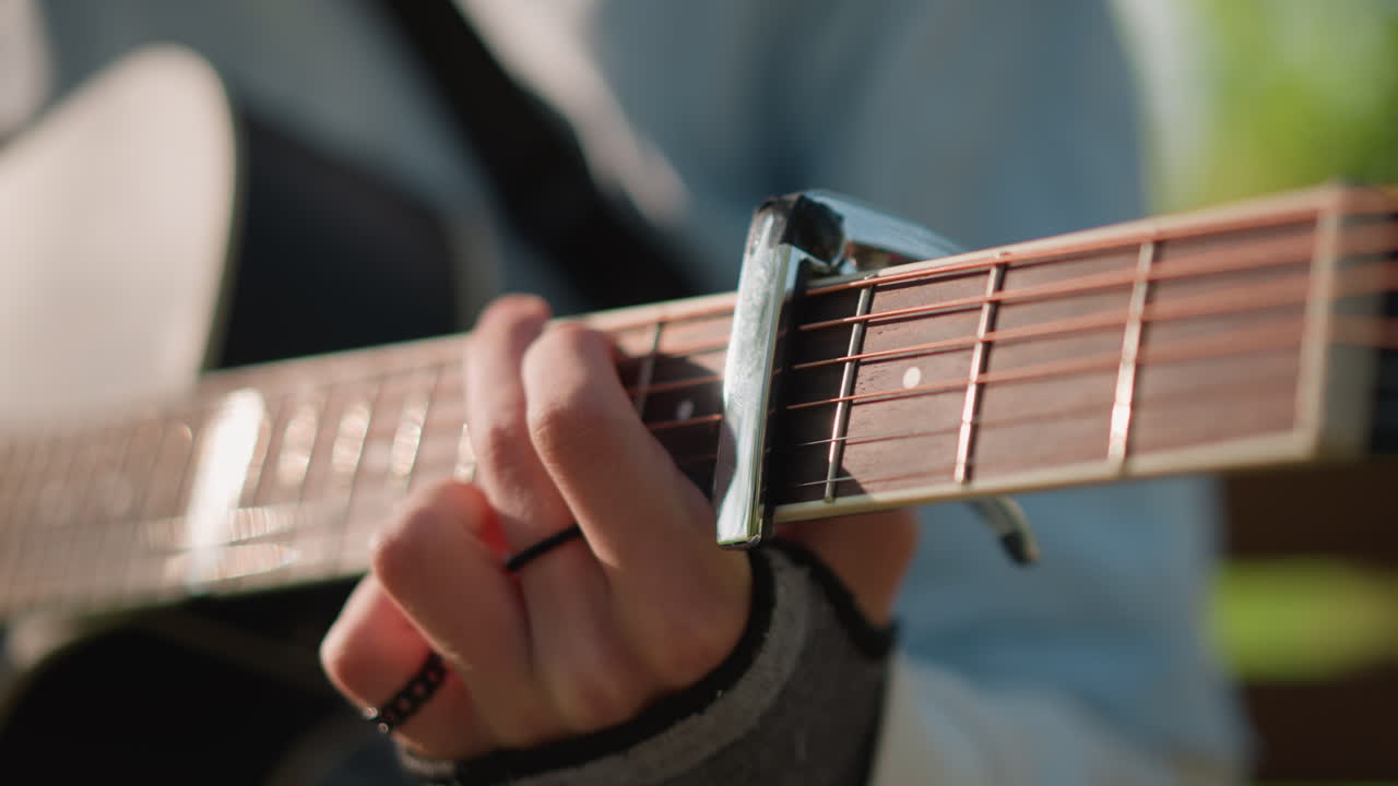 closeup acoustic guitar strings capo, fingerstyle busker composing in park, warm sunlight on hands, fingerless glove and ring, detailed fretboard motion, mellow indie melody, songwriter practicing