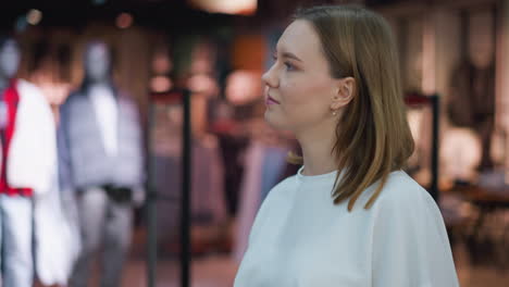 woman in white top gazing thoughtfully at clothing store display with mannequins in background, illuminated by warm lighting and surrounded by vibrant colors