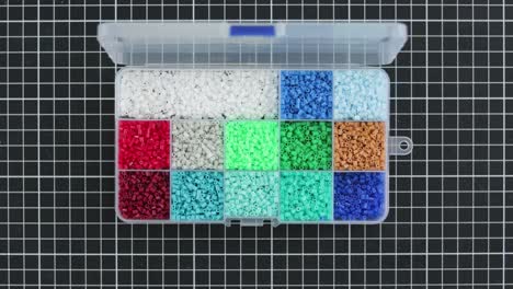 hands opening case filled with colorful mini fusion beads on grid background, overhead view