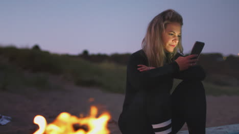 Frau-Sitzt-Auf-Einem-Surfbrett-Am-Lagerfeuer-Am-Strand-Und-Benutzt-Ihr-Mobiltelefon,-Während-Die-Sonne-Hinter-Ihr-Untergeht