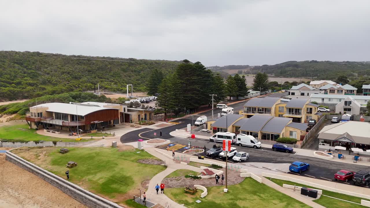 Drone footage captures Port Campbell's beachfront and town, highlighting buildings, greenery, and coastline under overcast skies