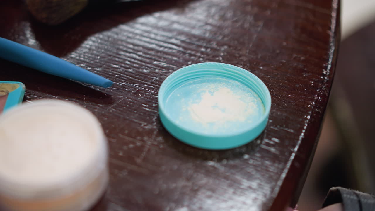 Close-up of lady hand holding purple makeup sponge, tapping into powder inside container, powder spills gently on surface, beauty routine setup with makeup tools on table in background