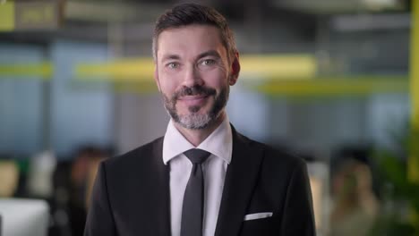 portrait of adult business man boss standing in office smiling looking at camera while employees are busy working in background
