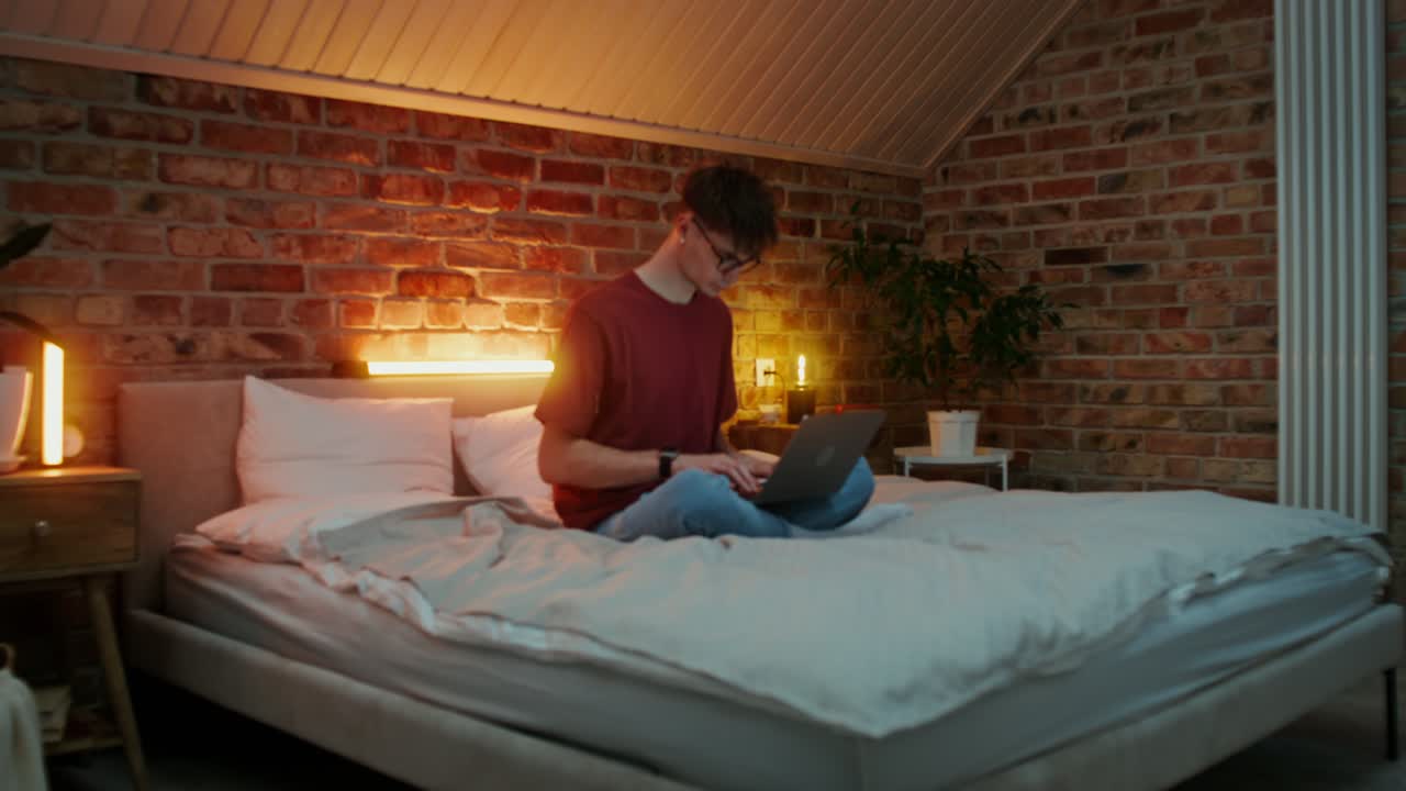 젊은 남자가 밤에 편안한 침실에서 노트북에서 일하고 있습니다.