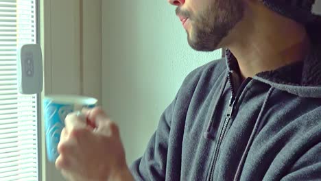 un joven mirando por la ventana y tomando café o té en casa.