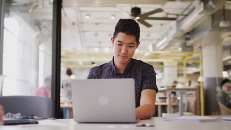 millennial asian man using laptop computer sitting at a table in an office meeting room, front view