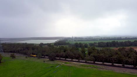 Vista-Aérea-Del-Tren-De-Carga-Amarillo-Que-Tira-De-Camiones-Hacia-La-Refinería-De-Alúmina-En-Un-Día-Lluvioso