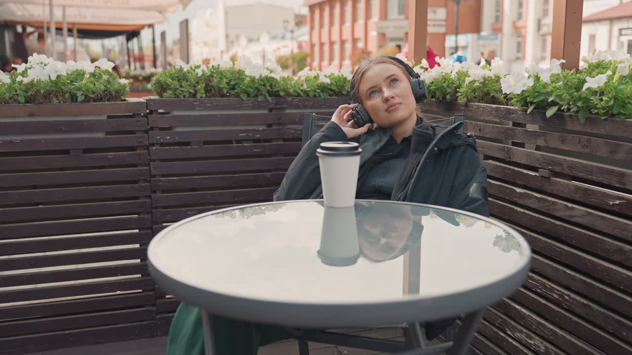 young woman with headphones at cafe, coffee cup on glass table, leaning back in chair, eyes closed then gazing upward, relaxed expression, puffer jacket, flower planter on wooden divider, urban street