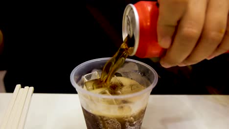 a hand pours dark soda from a red can into a clear plastic cup filled with ice, under warm indoor lighting, captured in a close-up static shot