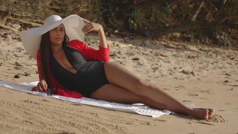 latijns model vrouw op het strand met een hoed op haar zij liggend zet een zonnebril op terwijl ze naar de camera kijkt