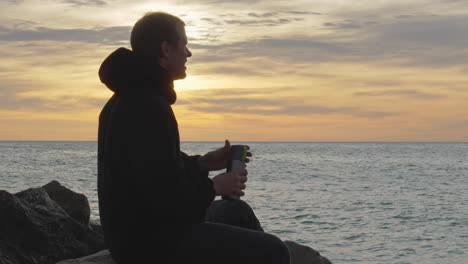 man enters, sits on a rock, opens thermos, pours in, drinks and enjoys the sea view during golden hour