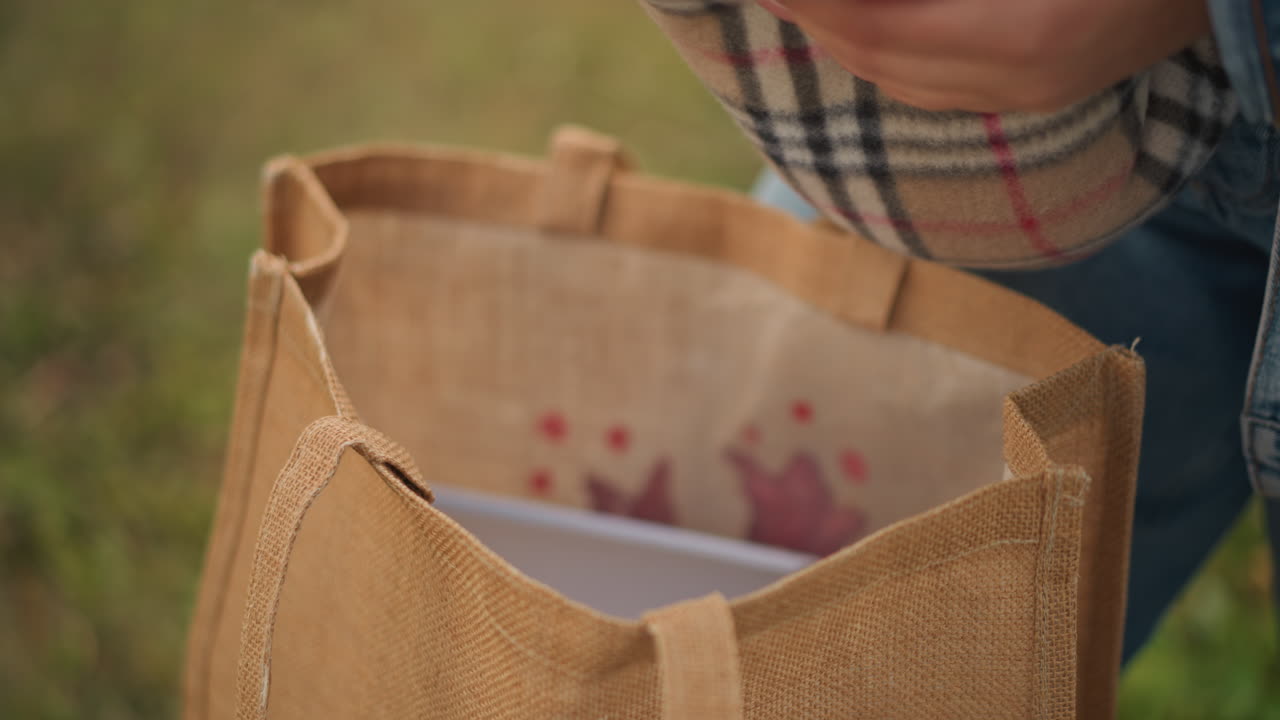 Mãos colocando cobertor xadrez em sacola de juta, close-up dos dedos dobrando o tecido e alisando o forro com padrão rosa, jeans e fundo de grama, detalhes táteis cuidadosos e vibração ecológica.