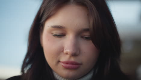 close-up of chic white woman with pink lips, subtle smile, focused on something in front of her, sporting a small nose ring, and a relaxed, thoughtful expression